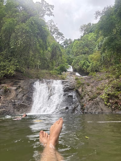 The image depicts a waterfall surrounded by lush green vegetation in Thai Mueang, Thailand. A person is submerged in the water at the base of the waterfall, with their feet visible above the water's surface. The waterfall cascades down a rocky cliff face. A rope hangs down into the water from the side. The sky is overcast.