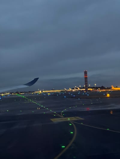 The image depicts an airport scene at night or late dusk, viewed from the window of an airplane on the ground. The sky in the upper left is dark and overcast. The primary subject is the illuminated airport tarmac or taxiway, which extends from the foreground towards the right side of the image.
The ground appears wet, reflecting the numerous artificial lights that define the airport infrastructure. Green lights are prominently displayed, outlining taxiway edges and guiding paths, with some forming curved lines in the mid-ground. Scattered blue lights are also visible, likely marking other sections of the airport. In the distance, yellow and orange lights illuminate various airport buildings and what appear to be parked aircraft.
A tall, distinctive tower with red and white horizontal stripes stands in the middle distance on the left side of the frame, emitting its own red warning lights. This is likely an air traffic control tower or a similar navigational structure. A portion of the airplane's wingtip, dark blue-grey, is visible in the lower left corner, establishing the passenger's perspective.
On the ground in the mid-right, a yellow rectangular marking with dark, indistinct text or symbols is visible, typical of airport signage. The overall atmosphere is that of a bustling yet quiet airport during non-daylight hours, with the wet surfaces adding a reflective quality to the scene. The location context for this active airport environment is Mitry-Mory, France.