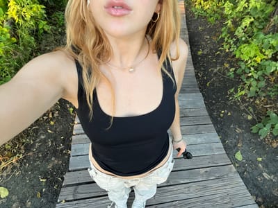 A selfie photograph taken from a low angle, looking up at a person standing on a wooden walkway. The person is wearing a black tank top, light-colored pants, and white sneakers. They are holding a small black object in their left hand. The background consists of green vegetation and trees, some of which can be identified as being of the region of Khwai, Botswana.