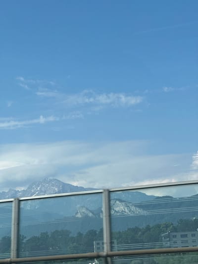 The image captures a scenic view through a glass pane, likely from a building. The foreground is dominated by a bright blue sky with scattered wispy clouds, suggesting fair weather. Reflected in the glass are towering mountains, some with patches of snow, indicating a mountainous region. Lush green trees are visible on the slopes of the mountains, and a building with multiple windows can be seen in the distance. The reflection also shows horizontal lines, possibly from the window frame or architectural features of the building where the photo was taken. The scene appears to be set during the daytime, with ample sunlight illuminating the landscape. The orientation of the image is sideways, making it appear as if the sky is on the left and the mountains are on the right. There is no visible text in the image.