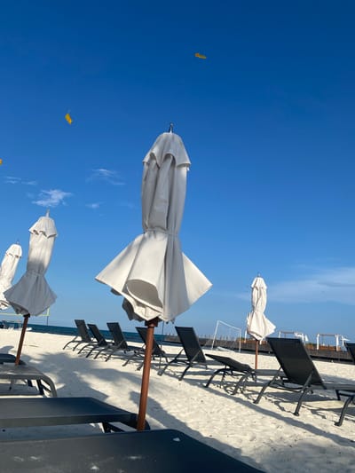 The image shows a beach scene in Marina Maroma, Mexico. The view is oriented towards the sky, which is a clear blue with scattered white clouds. Several closed white beach umbrellas are positioned at varying distances, interspersed among dark beach chairs on a sandy surface. In the distance, a soccer goal is visible on the beach. Some small yellow kites are scattered in the sky. The overall composition is bright and depicts a typical beach environment during daytime.