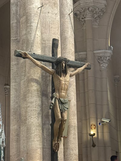 A horizontal view displays a sculpture of Jesus Christ on the cross, tilted slightly to the right. His body appears emaciated, wearing a simple garment. The cross is dark and rugged. The background is a textured wall of what seems to be light-colored stone or plaster, with architectural details such as cylindrical forms and ornate decorative elements. A security camera and a cross symbol with a light fixture are visible on the upper right. Thin wires are strung across the scene.