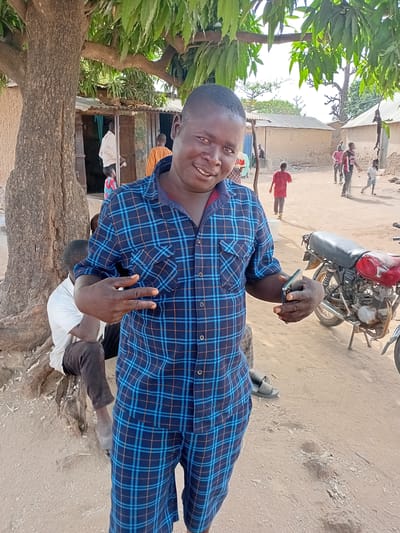 A man in a blue plaid shirt and pants stands outdoors near a large tree, holding a smartphone and making a peace sign. A red motorcycle is parked nearby. Several other people are visible in the background, some wearing red shirts. The scene appears to be in a rural area in Nata-Ala, Nigeria. The ground is sandy and there are simple buildings visible in the background.