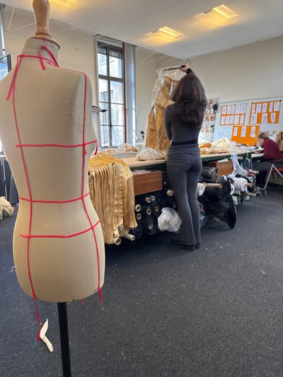 The image displays an indoor fashion design or textile workshop located in Arcueil, France. In the foreground, a cream-colored female dress form, marked with red ribbon strips indicating design lines, stands on a black pole base, positioned with its back towards the viewer. Behind it, a female individual, seen from the back, with long dark brown hair, wears a dark gray long-sleeved top and matching flared trousers. She is holding a golden-yellow garment, contained within a clear plastic garment bag, by its hanger, elevated above her head.
A large work table occupies the midground, featuring draped golden-yellow fabric, some exhibiting pleated textures. Beneath this table, numerous rolls of various fabrics, including black and white materials, are stored on shelving or directly on the dark gray carpeted floor. In the background right, a second individual with lighter hair, wearing a dark reddish-brown top, is seated at a table, concentrating on materials in front of them. The back wall features a display board adorned with multiple sketches or design drafts on white paper, some with orange backings. Large windows on the left wall provide natural light, revealing an exterior building with architectural details. Square, recessed ceiling lights are visible, and thin white strings are strung horizontally across the upper section of the room. The floor is covered with dark gray carpeting, showing scattered fibers. A small piece of white material rests on the floor near the base of the dress form.