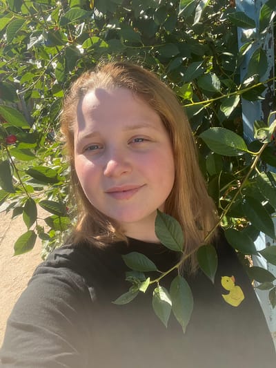 In Rakhinka, Russia, a young person with fair skin and blonde hair takes a selfie outdoors, surrounded by lush green foliage, possibly a cherry tree given the presence of small red fruits. The person is wearing a black t-shirt adorned with a yellow duck appliqué on the right side. Sunlight casts shadows and highlights on their face and clothing. In the background, a weathered blue structure, possibly a fence or post, is partially visible. The ground appears to be a light-colored, rough surface, perhaps concrete or stone. The overall scene is natural and sun-drenched.