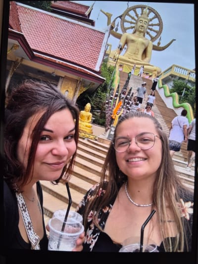 Two women are posing for a selfie. One woman has light brown hair and is wearing glasses and a white necklace. The other woman has dark brown hair and a crossbody bag with a patterned strap. Both women are holding drinks with straws. In the background is a large golden Buddha statue, a building with a red tiled roof, and a set of stairs. The setting does not appear to be in Mont-Saint-Martin, France, but seems like a Southeast Asian location. People are walking in the background.