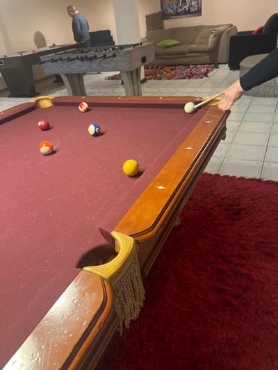 A pool table dominates the scene with red felt and a wooden frame. The cue ball is positioned near a side pocket, where a hand holds a pool cue. Several numbered balls are scattered on the table: the 8 ball, a yellow ball, a striped ball, and a red ball. A foosball table and a couch are visible in the background, along with a red shag rug.
