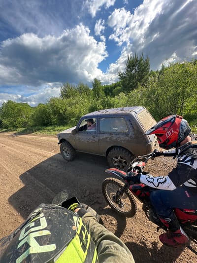 A photograph shows a muddy scene with a dirt road running through a wooded area under a cloudy sky. A blue, mud-covered SUV is parked on the road. A person wearing a red and black helmet and motocross gear is on a red and black dirt bike, positioned next to the SUV. Another person wearing similar motocross gear with a green and black helmet is partially visible in the lower right corner of the frame, suggesting they are also on a dirt bike. The location is Пепеляева, Russia.