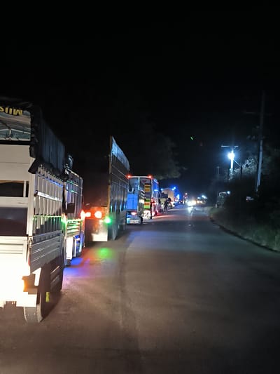 The image depicts a nighttime scene on a paved road in Lhoknga, Indonesia, dominated by a line of large trucks. In the immediate foreground on the left, a white truck features prominently, its side adorned with an array of colorful LED lights in green, blue, and orange, which reflect vibrantly on the dark road surface. A dark tarpaulin cover above its cargo area displays partially visible, inverted text that appears to read "MUSI."
Behind this truck, a series of other large trucks extends down the road, forming what appears to be a queue or a stationary convoy. Many of these trucks exhibit red taillights and various side marker lights. Further down the road, distinct blue lights are visible, possibly indicating emergency services, police, or simply more trucks with custom lighting. One truck in the mid-distance has "JIMU" visible as text on its front.
The setting is dark, with the black night sky suggesting late hours. Illumination comes primarily from the vehicles' lights and a bright street lamp positioned on the right side of the road. This lamp stands beside a utility pole and a dense line of trees and foliage, indicating a rural or semi-urban environment. In the distance, oncoming vehicle headlights are visible, adding to the artificial light sources. The activity suggests either a traffic hold-up, a checkpoint, or trucks parked along the roadside as part of a late-night transport operation. While no clear human figures are discernible in detail, faint shapes suggest people near the distant trucks. The overall scene is calm, with the colorful truck lights providing a striking contrast against the darkness.