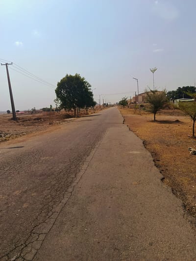The image depicts a straight, asphalt road stretching into the distance, possibly within the city of Rafin Pa, Nigeria. The road shows signs of wear and cracking, particularly on the left side. The roadside on both sides is characterized by dry, brown vegetation and sparse trees. A utility pole with power lines is visible on the left side of the road. Further down the road, street lights are spaced intermittently. Buildings can be seen in the distance on the right. The sky is a clear, pale blue. There are no people visible in the image.