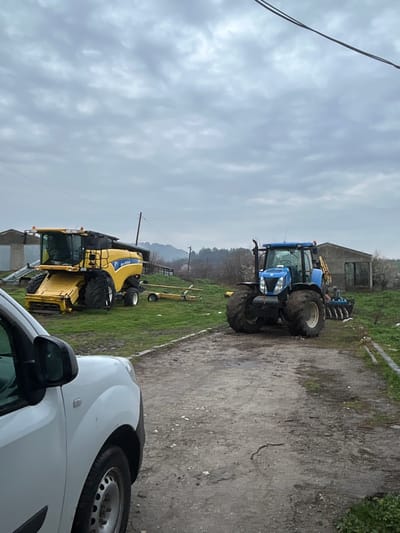 The image depicts an outdoor agricultural scene in Srednia, Bulgaria, under an overcast sky. In the foreground, the front left fender, wheel, and side mirror of a white vehicle (likely a van or car) are visible, facing right. A dirt and gravel path runs from the foreground towards the mid-ground.
To the left of the path, on a patch of green grass, a large yellow New Holland combine harvester is parked, oriented towards the left. Behind it, a yellow implement trailer is also visible. To the right of the path, a blue New Holland tractor, equipped with an attached implement (possibly a cultivator or disc harrow) at its rear, is parked, facing the viewer. The tractor's tires are visibly soiled.
In the background, on the left, a concrete or stucco building is partially visible. Further right, a larger, open-fronted barn or shed structure is present. The landscape features sparse vegetation, including trees and bushes, with distant hills visible under the cloudy horizon. Utility poles and overhead lines transect the upper portion of the scene. The ground is a mix of green grass and disturbed earth. No individuals are visible in the scene.