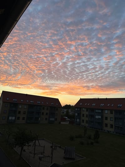 The image captures a sunset view from an elevated perspective, likely a balcony, overlooking an urban residential area. The sky is filled with a dramatic display of clouds, tinged with vibrant orange and pink hues from the setting sun, contrasting with patches of blue and gray. In the foreground, a dark, angled gutter runs along the top left edge of the frame. Below the sky are two identical apartment buildings with red tiled roofs and light-colored brick facades. The buildings have multiple windows and balconies, and appear to be part of a housing complex. In front of the buildings, a grassy area is visible, with a playground containing a sandpit, swings, and climbing structures. Trees are scattered across the lawn and along the edge of the property. The overall scene depicts a tranquil evening in a city setting. Given the information that the image is located in Roskilde, Denmark, the architecture of the buildings and the presence of a playground are consistent with typical residential areas in such a location. There are no people visible in the image.