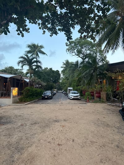 The image depicts a scene in Ban Bang Niang, Thailand. In the foreground, a sandy, unpaved road leads into the distance. On the left side of the road, there is a building with a wooden exterior, a white awning, and outdoor seating. A sign with illuminated text, possibly for a restaurant or shop, is visible on a concrete base. Further down the road on the left, more buildings with similar architectural styles are present, nestled amongst lush vegetation.
On the right side of the road, a modern building with dark siding and large windows is visible, with potted plants and a red umbrella outside. A white car is parked on the right side of the road, and a black car is parked on the left side further down. Several motorcycles are also parked along the side of the road.
The background consists of dense tropical foliage, including palm trees and other trees, under a cloudy sky. The overall atmosphere is that of a tropical coastal town with a mix of natural beauty and established commercial establishments. The lighting suggests it is either early morning or late afternoon, with soft, diffused light. There are no people clearly visible in the image.