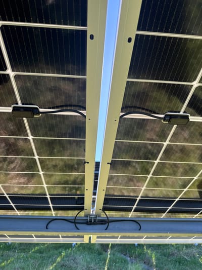The image provides an upward-facing view of the underside of a ground-mounted photovoltaic solar panel array, located in Eastleigh, United Kingdom. The scene is brightly lit by natural sunlight, indicating a clear sky. In the upper two-thirds of the frame, the dark, grid-patterned undersides of multiple solar panels are visible, with their individual cells delineated by thin white lines. A distinct, bright vertical strip of clear blue sky bisects the upper central portion of the image, running between two rows of solar panels. This gap is flanked by light-colored metallic mounting rails that support the panels.
Black electrical cables are evident across the scene. In the upper left and right, individual cables connect to black, rectangular electrical optimizers or junction boxes securely mounted to the underside of the solar panels. These cables loop downwards. Towards the bottom center, several black cables converge, are neatly bundled, and secured by clips to a robust, horizontal metallic support beam that forms part of the array's structural framework. A central electrical connector is visible at this convergence point on the beam. Below this metallic structure, green grass is clearly visible, confirming an outdoor installation in a grassy area. No human figures are present.