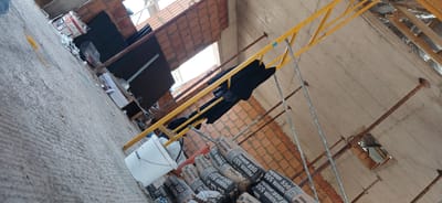 The image depicts an indoor construction site in Nimoreni, Moldova. The scene is a partially constructed building with exposed brick walls and a concrete floor. A yellow scaffolding structure is present, with dark clothing draped over it. A white bucket and several bags of cement labeled "PREMIUM 550" are visible on the floor. In the background, there are glimpses of roofing materials and additional construction supports. The overall impression is one of an active but untidy worksite.