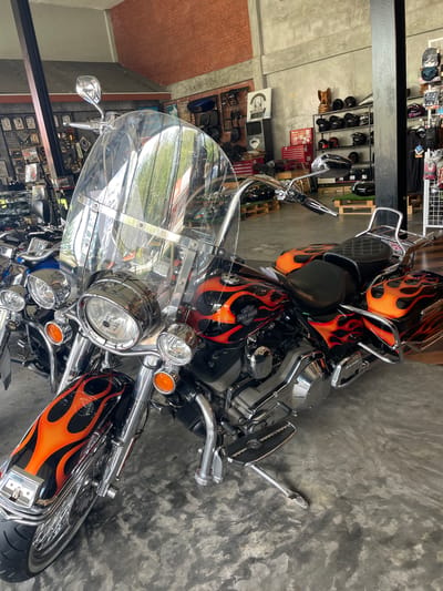 The image depicts the interior of a motorcycle shop located in Ban Yid, Thailand. A black Harley-Davidson motorcycle with prominent orange and red flame graphics dominates the foreground and center. This motorcycle features a large, clear windshield, chrome handlebars with integrated mirrors, a large central headlight flanked by two smaller chrome lights, and amber turn signals. The front fender is black with flame graphics, and numerous chrome components are visible, including the engine casing, exhaust pipes, and footboards. The motorcycle has a two-piece seat, with a smooth black front seat and a diamond-stitched black rear seat, accompanied by matching flame-painted saddlebags and a chrome luggage rack at the rear. The fuel tank displays "HARLEY-DAVIDSON COMPANY" text.

To the left, partially visible behind the main motorcycle, is a blue motorcycle. The shop's floor is composed of grey polished concrete with darker, irregular patterns. The walls vary, with exposed red brick on the left side and grey concrete or stucco on the rear and right walls. In the background, a black metal shelving unit displays multiple motorcycle helmets, predominantly black, with one featuring orange accents. A wooden elephant statue rests atop this shelving unit. A red multi-drawer toolbox is visible to the left of the helmet display. Various merchandise racks, displaying small items and potentially clothing or parts, are positioned along the left and right walls. Wooden pallets are scattered on the floor in the mid-ground. No individuals are present in the scene.