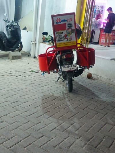A Honda motorcycle with license plate DM 2247 RO is parked on a herringbone-patterned brick sidewalk in front of a building with a light-colored facade. Attached to the rear of the motorcycle are two red cylindrical baskets. A sign above the baskets advertises "Alfgift". Behind the motorcycle, two individuals are visible inside a store with shelves displaying various items. One individual, wearing a red headscarf, is interacting with another person who is wearing a black t-shirt and shorts. A coconut is resting on the sidewalk to the right of the motorcycle. The scene appears to be in a commercial area in Tenggela, Indonesia.