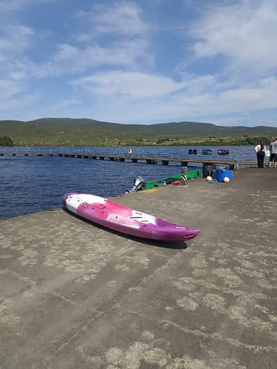 A pink and white kayak rests on a concrete pier beside a body of water. In the background, a long wooden pier with blue railings stretches across the water towards a green, hilly landscape. The sky above is blue with scattered white clouds. Two people, a woman and a man, stand at the end of the wooden pier, facing the water. Several other boats and buoys are visible on the concrete pier. The scene appears to be in Loughanure, Ireland, likely a marina or watersports facility given the presence of kayaks and a pier.