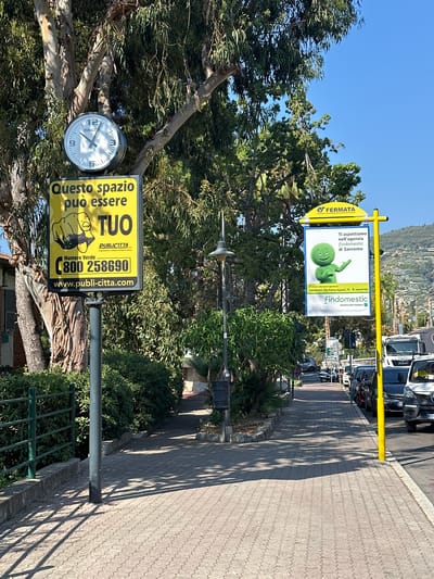 This image depicts a street scene in Ospedaletti, Italy, under a clear blue sky. On the left side of the frame, a clock is mounted on a pole, positioned below a large tree with visible foliage. Next to the clock, a yellow advertisement displays an illustration of a pointing finger and the text "Questo spazio può essere TUO" (This space can be yours), along with a green phone number "800 258690" and a website address "www.publi-citta.com".
Further down the street, on the right, a yellow bus stop sign with "FERMATA" at the top is visible. Beneath it, another advertisement features a green cartoon character and the text "Ti aspettiamo nell'agenzia Findomestic di Sanremo" (We are waiting for you at the Findomestic agency in Sanremo). This ad also includes an address and a phone number.
The street is paved with interlocking bricks, and shadows from trees and overhead structures are cast across it. A row of parked cars lines the right side of the street. In the background, lush green vegetation and trees are prominent, with a hillside dotted with buildings visible in the distance. The overall environment appears to be a sunny, urban setting.
