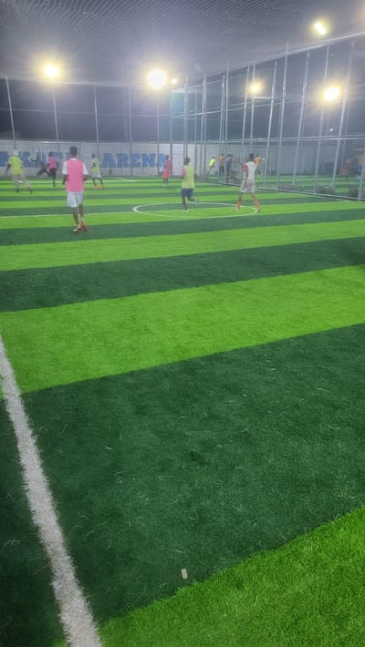An artificial turf football or futsal pitch in Babawa, Nigeria, is depicted at night, brightly illuminated by multiple overhead floodlights. The playing surface features alternating stripes of light and dark green artificial grass, with visible white lines delineating the field boundaries and a central circle. The entire playing area is enclosed by tall metal fences and netting, with a larger net spanning overhead. In the background, a light-colored wall displays partial blue lettering, including "ARENA." Approximately 10 to 15 individuals, primarily males, are present on the pitch, actively engaged in a game. They are dressed in casual sportswear, with some wearing colored bibs or vests in pink, yellow, and orange. One individual in a pink vest and white shorts is prominently positioned in the foreground, facing away from the camera. Other players are scattered across the field, in various postures indicating running and active play. No ball is distinctly visible.