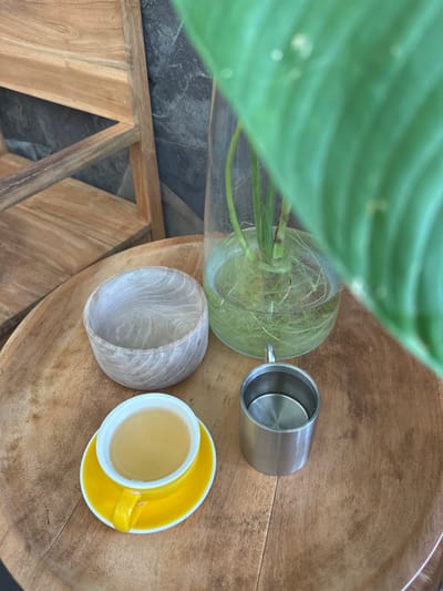 A high-angle shot shows a wooden table with a cup of tea, a metal cup, and a plant in a vase in Licin, Indonesia. The wooden table has a rounded, bowl-shaped top. A green leaf dominates the left side of the frame, partially obscuring a glass vase filled with water and roots. To the right of the vase is a stainless steel cup lying on its side. A small, wooden bowl sits to the left of a bright yellow cup and saucer filled with a pale liquid, presumably tea. The background features a glimpse of tiled flooring and wooden support beams. The scene is well-lit, highlighting the textures of the wood and the reflective surfaces of the glass and metal.