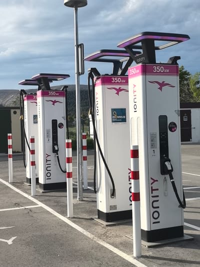 This image captures a row of four high-power electric vehicle (EV) charging stations, branded "IONITY," situated in a parking lot in Sallent, Spain. Each charger is a prominent white pillar with magenta accents, featuring "350 kW" clearly displayed, indicating its fast-charging capability. A distinctive magenta bird logo, possibly a swallow or swift, is visible on the upper panel of each unit, which also includes an illuminated top section. Black charging cables are coiled or hanging from the side of each station.
The charging bays are delineated by red and white striped bollards, presumably for vehicle protection, and the asphalt parking surface features white parking lines and a painted directional arrow. A tall light pole stands among the chargers. In the background, there is a simple utility building with a dark door, and distant hills or mountains are visible under a partly cloudy sky, suggesting a somewhat rural or industrial setting.
The scene is set during the daytime, likely late afternoon, given the natural light and visible shadows. No people or active charging events are taking place. Visible text includes "IONITY" vertically on the side of the chargers and horizontally at the top. Charger numbers "1" and "2" are discernible on two of the units. One charger bears a "moves III" sticker with "Mobilised Efficace & Sostenible," and another shows "IONITY Passport."