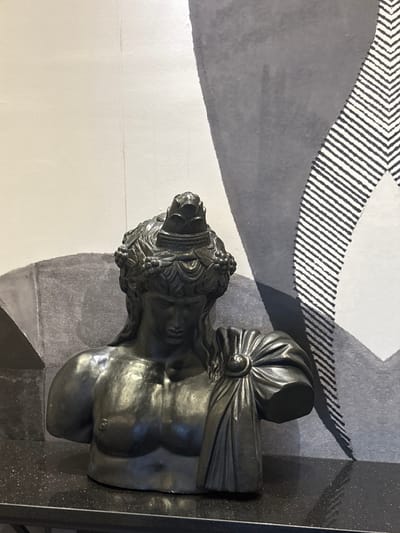 A dark, metallic bust of a male figure with long hair and a laurel wreath rests on a black, speckled surface. The figure is depicted from the chest up, with defined musculature and draped fabric across the chest. The background is a light-colored wall with a large, abstract mural. The mural features a grayscale design with geometric shapes and a pattern of diagonal black lines. The bust is positioned centrally in the frame, with the mural occupying the majority of the background space. The lighting is even, casting soft shadows. There are no people or animals visible in the image. This scene is located in the city of Khirbet es-Souk, Jordan.