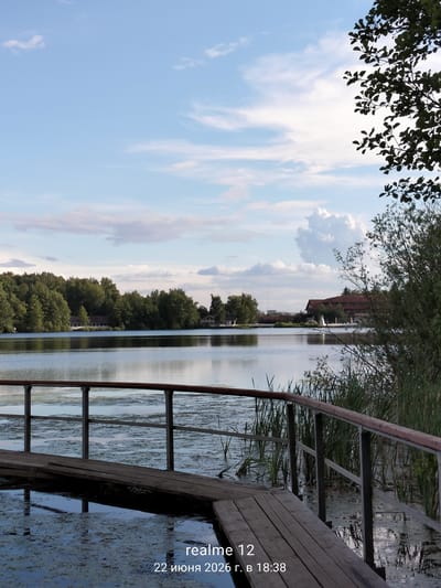 The image shows a tranquil lake scene with a wooden boardwalk in the foreground. Lush green trees line the opposite bank of the lake, with buildings visible amongst them. The sky is partly cloudy, with a mix of blue sky and white clouds. The water is calm, reflecting the sky and surrounding trees. The wooden boardwalk, made of planks, extends into the water and is bordered by a metal railing with wooden accents. Reeds and other aquatic vegetation are present along the water's edge and in the water near the boardwalk. The overall impression is of a peaceful natural environment, possibly a recreational area. The timestamp indicates the image was taken on June 22, 2026, at 18:38 in Авиатор Снт, Russia. There are no people or animals visible in the image.