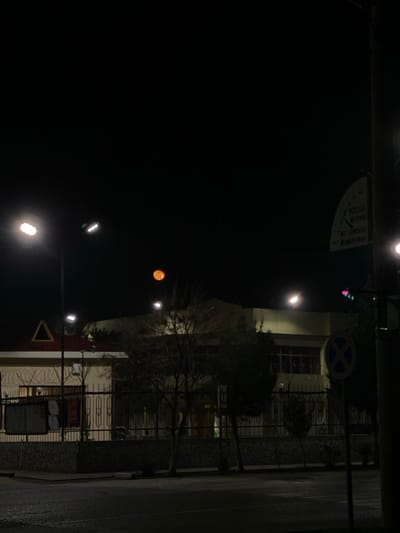 The image depicts a nighttime street scene in Karmana, Uzbekistan. The dominant feature in the upper-left sky is a distinct, orange-hued moon. The foreground consists of a dark asphalt road, potentially wet, bordered by a sidewalk and a low stone wall. In the midground, two buildings are visible behind a dark metal fence with a stone base. The building on the left is a single-story structure with a light-colored facade and a red sloped roof featuring a triangular apex; barbed wire tops its fence. A notice board is positioned near the fence of this property. To the right, a larger, two-story building with a lighter facade and a flat roof stands further back. Leafless trees and shrubs are scattered in front of and between the buildings. Multiple streetlights illuminate the scene with bright white light. On the far right, a utility pole supports a blue circular "No Stopping" sign and a curved white informational sign with black text in Uzbek. A traffic light with a green signal is partially visible on the same pole. No people, discernible actions, or interactions are visible.