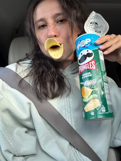 A close-up, tilted shot captures a person inside what appears to be a car, engaged in a playful moment with Pringles. The individual has dark, wavy hair and visible freckles on their nose, with a single Pringles chip playfully positioned in their mouth. Their right hand, adorned with a ring and manicured nails, holds an open green Pringles can.
The can clearly displays the "PRINGLES" logo and the flavor "CHEESE & ONION," along with illustrations of cheese and onions. The blue, peeled-back lid features a "POP PLAY" logo and a "SCAN" prompt with a QR code. Visible text on the can includes nutritional information (e.g., "100g: 2185 kJ/523 kcal") and manufacturer details such as "Pringles BVBA SPRL, Eggestraat 1, 2000 Mechelen," along with multilingual indications of origin including "Belgia / Бельгия."
A grey seatbelt diagonally crosses the person's torso and light-colored clothing, confirming the setting as a vehicle. The background is indistinct, showing parts of the car's interior. The overall scene suggests a casual and lighthearted snack break.