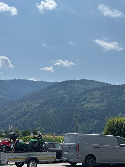 This image depicts a roadside scene with a mountainous backdrop, likely near Pusarnitz, Austria. In the foreground, a trailer is loaded with two motorcycles: a red one on the left and a green one on the right. The green motorcycle is a sport-touring bike. A gray panel van is parked to the right of the trailer, and a dark-colored car is partially visible between the trailer and the van. Behind the vehicles, a field of crops, possibly corn, stretches towards a dense, forested mountain range that dominates the background. The sky is blue with scattered white clouds.
