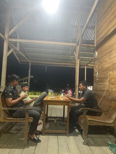 The media file shows three individuals gathered at an outdoor, covered seating area, likely a cafe or restaurant, in Cotkruet, Indonesia. The scene is illuminated by artificial lighting, suggesting it is nighttime. The structure appears to be built with wood and corrugated metal, giving it a rustic, open-air ambiance.

Two of the individuals are seated at a wooden table, engaged in conversation. One person, wearing a dark t-shirt and dark pants, is leaning forward towards the table, seemingly interacting with something on it. Another individual, wearing a light-colored t-shirt and dark pants, is also seated at the table, looking away from the camera. The third person is seated on a wooden bench, facing away from the camera, with their legs crossed.

On the table, there are a few items, including what appears to be glasses, a bottle, and possibly some food or snacks. In the background, a red motorcycle is visible, suggesting that transportation is readily available in the area. The overall mood appears relaxed and informal, with the individuals enjoying each other's company. The weather appears to be clear, as there are no visible signs of rain or adverse conditions. The location cues, such as the wooden structures and the presence of a motorcycle, are consistent with a setting in Indonesia. There is no visible text in the media file.
