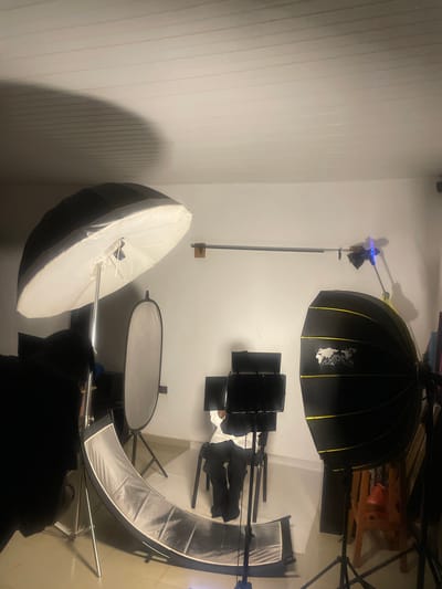 The image depicts an indoor studio setup, likely for photography or videography. The scene is illuminated by several studio lights, including a large umbrella softbox on the left, a rectangular reflector in the middle, and a large parabolic softbox on the right. A person, dressed in dark clothing, is seated on a chair in the center of the frame, facing away from the camera, with their head turned to the right. They appear to be looking at a setup of three monitors positioned on stands in front of them.
The room has white walls and a white slatted ceiling, with shadows cast by the lighting equipment. A metal rod extends horizontally across the wall near the ceiling, supporting one of the lights. Various other studio equipment, including stands and possibly fabric backdrops, are visible. The floor appears to be tiled.
There are no discernible landmarks or features that would indicate the specific location within South Ropp, Nigeria. The scene is sterile and functional, focused entirely on the studio equipment and the seated individual.