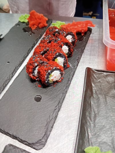 The image shows a close-up view of a sushi platter. The sushi rolls are topped with red roe and appear to be made with black rice. They are arranged on a dark, slate-like platter alongside pickled ginger and wasabi. Another dark plate is visible to the right, and a clear plastic container with red sauce is in the background. The scene is set on a metallic surface, likely a table in a restaurant or food preparation area. The lighting suggests an indoor setting. Without further context, it is impossible to determine the specific location within Mykolaiv, Ukraine, or to identify any individuals present. The image focuses solely on the presentation of the sushi.