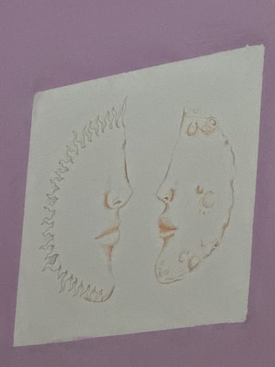 The image shows a drawing on a piece of paper. The paper is off-white and has two separate drawings on it, one above the other. The top drawing is a somewhat amorphous shape, with several circular features and what appears to be a profile of a face. The bottom drawing is a half-moon shape with jagged edges. Both drawings are rendered in a light brown color. The paper is against a lavender or light purple background. There is no indication of a specific location in Castelnuovo di Porto or any indication of people, subjects, objects, actions, or interactions, beyond the drawings on the paper and the background.
