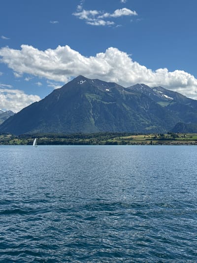 A wide-angle vertical shot captures a vast blue lake in the foreground, with choppy waves reflecting the bright sunlight. In the midground, a small white sailboat with a single sail glides across the water. Beyond the lake, a lush green landscape rises, dotted with trees and homes, leading to the base of a large, dark green mountain. Patches of snow are visible on the upper slopes of the mountain, which is partially obscured by large, fluffy white clouds against a brilliant blue sky. The scene depicts a serene natural landscape.