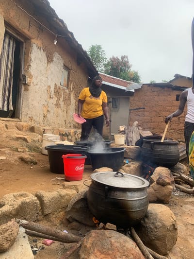 In Zhwarki Mile 10, Nigeria, an outdoor cooking scene unfolds. A woman wearing a yellow t-shirt and a black hijab is standing next to several large cooking pots. She holds a pink sieve. Steam rises from at least one of the pots, indicating cooking is in progress. Several large black plastic tubs are also present, some filled with water or ingredients. A red bucket with the label "Royco" is visible. The cooking area appears to be set up outdoors with large rocks used as a makeshift stove for at least one of the pots, where a fire is burning. Behind the cooking area, a traditional mud-brick house with a corrugated metal roof stands. The ground is dirt, and scattered stones and debris are present. Another person, partially visible on the right, is also tending to the cooking with a wooden spoon. The sky is overcast.