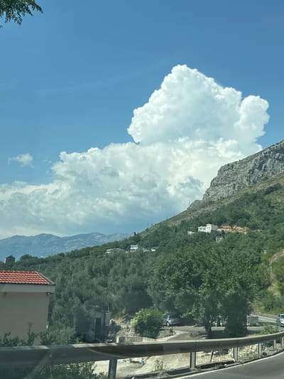 The image captures a scenic view in Rijeka Reževići, Montenegro, under a bright blue sky dotted with large, fluffy white clouds. A steep, rocky mountainside covered in lush green vegetation dominates the right side of the frame. Several white buildings are nestled on the slope, indicating a residential area. In the foreground, a building with a red-tiled roof is partially visible on the left. A road with a metal guardrail runs along the bottom of the image, with a few vehicles visible on or near the road. The overall impression is a serene, natural landscape with human habitation integrated into the mountainous terrain.