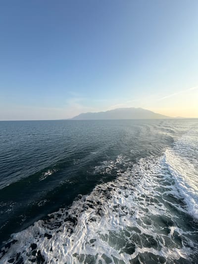 The image displays a panoramic seascape near Kato Karioutes, Greece, observed from a moving vessel. The lower portion features a deep blue-green body of water, prominently marked by a wide, white, turbulent wake extending diagonally from the lower right towards the lower left. Ripples are visible across the water's surface to the left of the wake. A distant, low-lying mountain range or large island stretches across the horizon from the mid-left to the far right, appearing hazy due to distance. The sky above is clear, transitioning from a light blue near the horizon to a deeper blue at the top, with a subtle yellowish tint on the far right horizon. A faint linear cloud or contrail is present in the upper right sky. No individuals or other discernible man-made objects are visible within the frame.