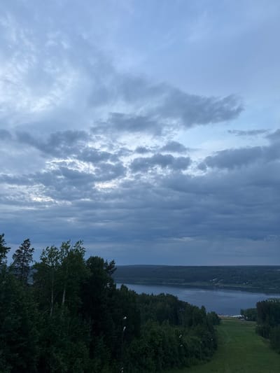 The image displays an expansive natural landscape under overcast skies, viewed from an elevated vantage point in Korobitsyno, Russia. Approximately two-thirds of the frame is occupied by a dense, multi-layered cloud system, predominantly grey and muted blue, with variations in density and some areas of diffused brighter light. The lower third features a dense, dark green temperate forest, comprising both coniferous and deciduous trees. A broad, calm body of water, reflecting the grey-blue sky, traverses the midground horizontally, separating the foreground forest from the distant landscape. On the right side of the midground, a cleared grassy slope, consistent with a ski run, descends towards the water, flanked by dark green foliage and marked by several vertically oriented light poles. The far bank of the water is lined with an unbroken expanse of forest, with indistinct structures or landforms barely discernible on the distant horizon. No individuals, specific actions, or other discernible objects are present.