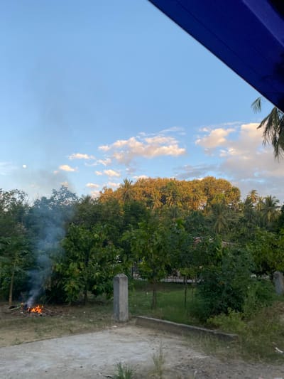The image shows a landscape in Payakumbuh, Indonesia. The sky is blue with scattered white clouds. There is lush green vegetation, including trees and bushes. A fire is burning on the ground, producing smoke that rises into the air. There's a concrete pathway or structure on the right. A part of a blue structure is visible in the top left corner. The moon is also visible in the sky.