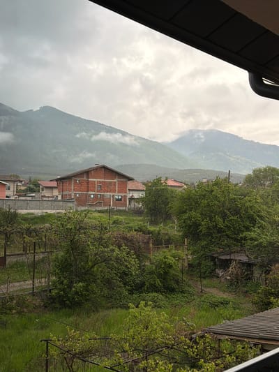 The image depicts a mountainous landscape near Resilovo, Bulgaria, under overcast skies. A partially constructed brick building is visible in the foreground, with a mountain range rising in the background, partly obscured by mist. Vegetation is prominent, with trees and grass covering the hillside. Adjacent to the unfinished brick structure, other residential buildings are visible. Power lines stretch across the frame, and elements of architecture are visible at the top and right of the frame.