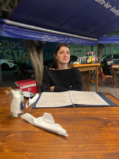The image depicts a young woman sitting at a wooden table in an outdoor dining area. She is positioned next to a menu and a container with napkins. A red delivery box is also on the table. The restaurant appears to be in Dock Sud, Argentina, indicated by a blue awning with the word "Dock" visible. The background features a bar with various bottles and a delivery motorcycle.