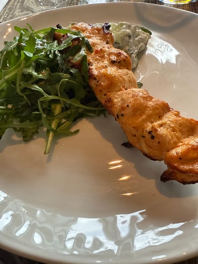 A white, irregularly shaped ceramic plate presents a single grilled chicken skewer, oriented diagonally from the upper right to the lower left. The skewer comprises several pieces of light brown, seared chicken, visibly seasoned with dark specks. To the left of the chicken skewer, a mound of fresh green arugula is positioned, appearing lightly dressed. Behind the upper section of the skewer, a portion of a pale green or off-white creamy sauce, possibly tzatziki, is visible. The plate's surface exhibits multiple reflections of ambient light, suggesting an indoor environment, potentially a restaurant, with indistinct details reflected from above. No individuals, explicit actions, or interactions are depicted. The image itself contains no discernible visual markers to confirm the specified location of Felixstowe, United Kingdom.