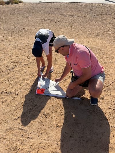 In Antigua, Spain, an outdoor scene depicts two individuals on dry, sandy ground under bright sunlight. A younger individual, wearing a black baseball cap, white t-shirt with a dark graphic, dark shorts, and white flip-flops, is bent over and focused on a kite lying on the ground. An adult male, wearing a beige baseball cap, sunglasses, a pink polo shirt, striped shorts, and dark blue shoes, is kneeling beside him and also focused on the kite. Both individuals appear to be engaged in the assembly or preparation of the kite, which is white, grey, and orange, bearing the text "VODAFONE MERCEDES." Distinct, elongated shadows of the two figures are cast on the light brown, uneven ground, which shows minimal sparse vegetation in the distant background, alongside a paved surface.