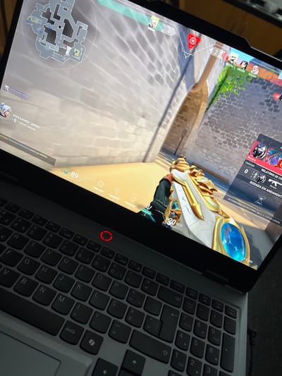 The image displays a laptop screen showing the first-person perspective of the video game Valorant. A gold and white weapon is visible, with the in-game HUD displaying health, map, and player information. The laptop's keyboard is visible to the right of the screen, featuring standard QWERTY layout and function keys. A red power indicator light is lit on the laptop's chassis. The overall scene captures a moment of gameplay on the laptop.