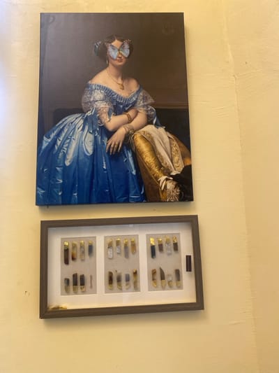 The image shows two framed pieces of art on a yellow wall. The top frame contains a painting of a woman in a blue dress sitting in a chair, with a blue butterfly covering her eyes. The bottom frame contains a display of small, rectangular objects with gold trim. The objects are arranged in three rows of six. The objects appear to be pieces of jewelry. The location based on the coordinates provided is in Buea, Cameroon. There are no people visible in the image. The image appears to be taken indoors, with no evidence of natural light. The image is taken at an angle.