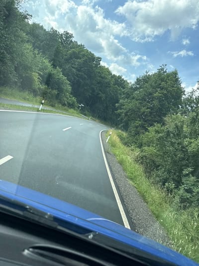 A car-mounted, eye-level, medium shot captures a view of a paved, winding road cutting through a dense forest. The road, with white lane markings, curves to the right, disappearing into the greenery. The forest, comprised of various shades of green trees, lines both sides of the road, with a strip of tall grass and gravel bordering the right side of the asphalt. The sky above is a bright blue, dotted with fluffy white clouds. A small section of the blue car's hood is visible in the foreground. The scene suggests a drive through a rural, wooded area.