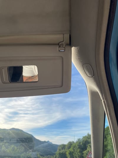 The image shows the interior of a car, looking out through the passenger side window. In the foreground, the beige sun visor is visible, with a mirror on its underside reflecting part of a person's torso with tattoos. To the right of the sun visor, a gray pillar with "AIRBAG" written on it is visible. Through the window, a bright blue sky with scattered white clouds is seen above rolling green hills and trees. The overall scene suggests a sunny day while driving through a rural or mountainous area. The location is specified as Obzovica, Montenegro.