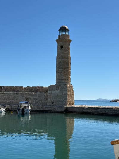The image depicts the Venetian harbor of Rethymno, Greece, on a clear, sunny day. A tall, stone lighthouse with a lantern room at its peak stands prominently on a fortified wall extending into the turquoise waters of the harbor. The lighthouse is constructed of rough-hewn stone blocks, with a circular opening visible on its shaft. A section of the ancient stone wall, partially in ruin, stretches horizontally, with a person walking along its top. In the foreground, calm blue water reflects the clear sky and the stone structures. Two small motorboats are docked near the wall, one of which is partially visible on the left. Further out in the distance, a second, smaller lighthouse can be seen on a breakwater. The overall scene conveys a sense of historical significance and coastal serenity.